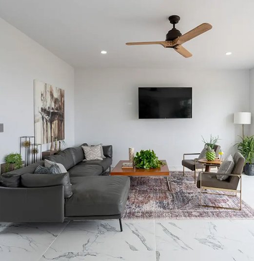 Un primer plano de la sala de estar moderna en una casa. La habitación tiene paredes blancas, suelo de mármol blanco veteado, un sofá seccional de cuero gris oscuro, una mesa de centro de madera oscura y una alfombra estampada. Hay una televisión de pantalla plana montada en la pared y un ventilador de techo de madera. Varias plantas de interior y obras de arte decoran el espacio.