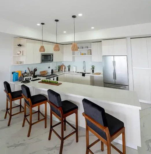  Una toma de cerca de una cocina moderna y luminosa. La cocina tiene gabinetes blancos, encimeras blancas, un protector contra salpicaduras de vidrio celeste y electrodomésticos de acero inoxidable, incluyendo una nevera y un microondas. En el centro hay una isla grande con cuatro taburetes de bar de terciopelo negro y madera en el primer plano. El suelo es de mármol blanco veteado.